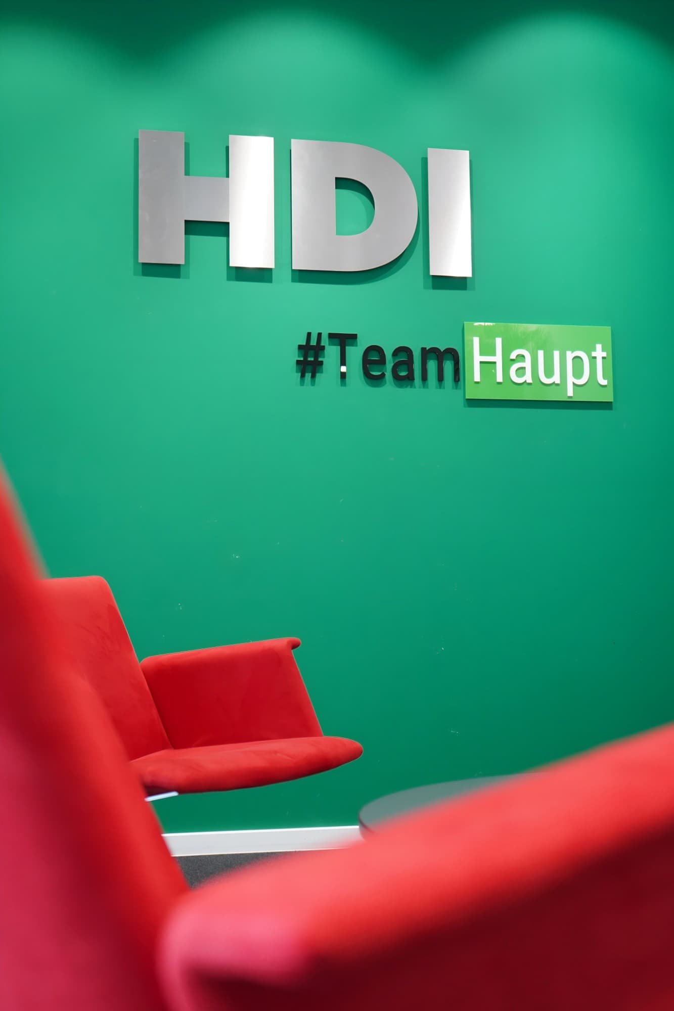 Wartebereich der Agentur: rote Sessel vor der grünen Wand mit HDI-Schriftzug und #TeamHaupt